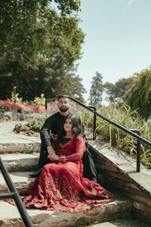A couple poses on stone steps outdoors with the person in a flowing red lace dress and another in black attire.