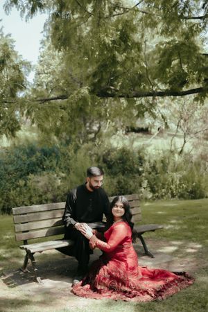 A couple in contrasting outfits sits together on a park bench surrounded by greenery.