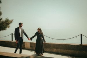 A couple holds hands while standing on a waterfront walkway with a railing.