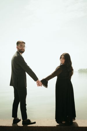A couple in formal black attire holds hands while standing by a misty waterfront.
