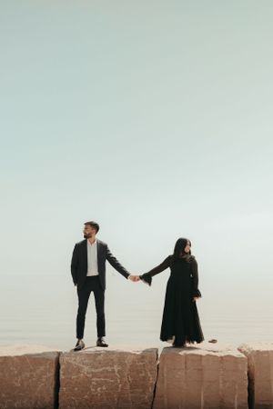 A couple in black attire holds hands while standing on stone blocks against a light sky.
