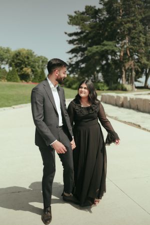 A couple takes a romantic stroll on a concrete path near a waterfront park.