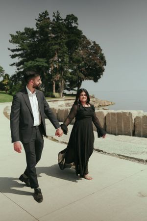 A couple walks hand in hand along a waterfront path with trees in the background.