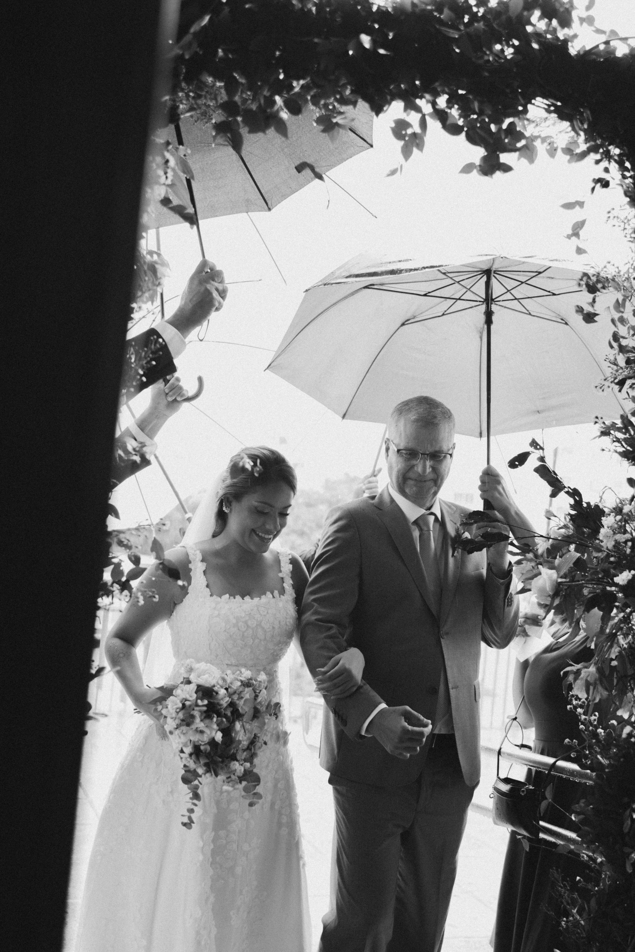 A black and white wedding photo showing a couple under an umbrella framed by foliage.