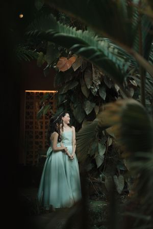 Two figures in matching mint green dresses stand among lush tropical foliage in a garden setting at dusk.