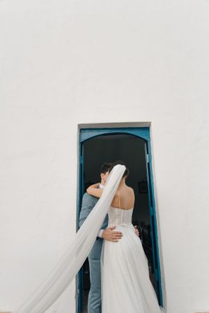 Newlyweds embrace in a blue doorframe with flowing white veil and gray suit.