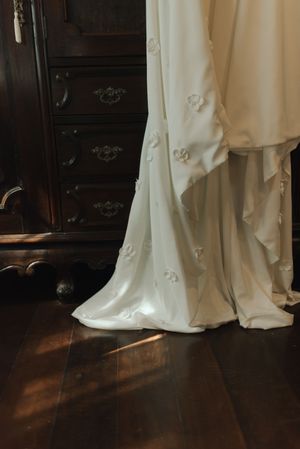 A white wedding dress with delicate floral details flows onto dark hardwood flooring beside an antique dresser.