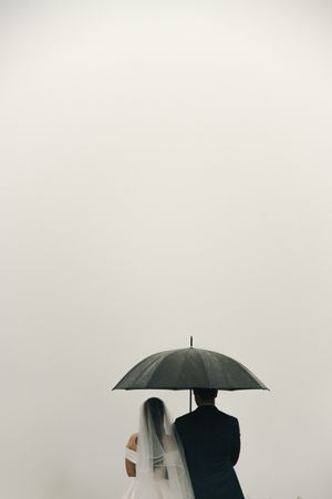 Minimalist black and white photo of couple sharing black umbrella against light background, with bride wearing flowing veil.