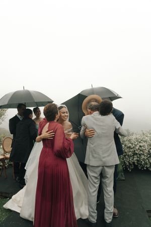 Wedding party members huddle under umbrellas on a rainy day with misty backdrop.