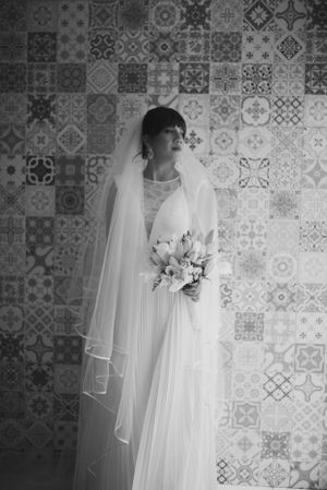 Bride in elegant white gown and veil poses against patterned tile wall with bouquet.
