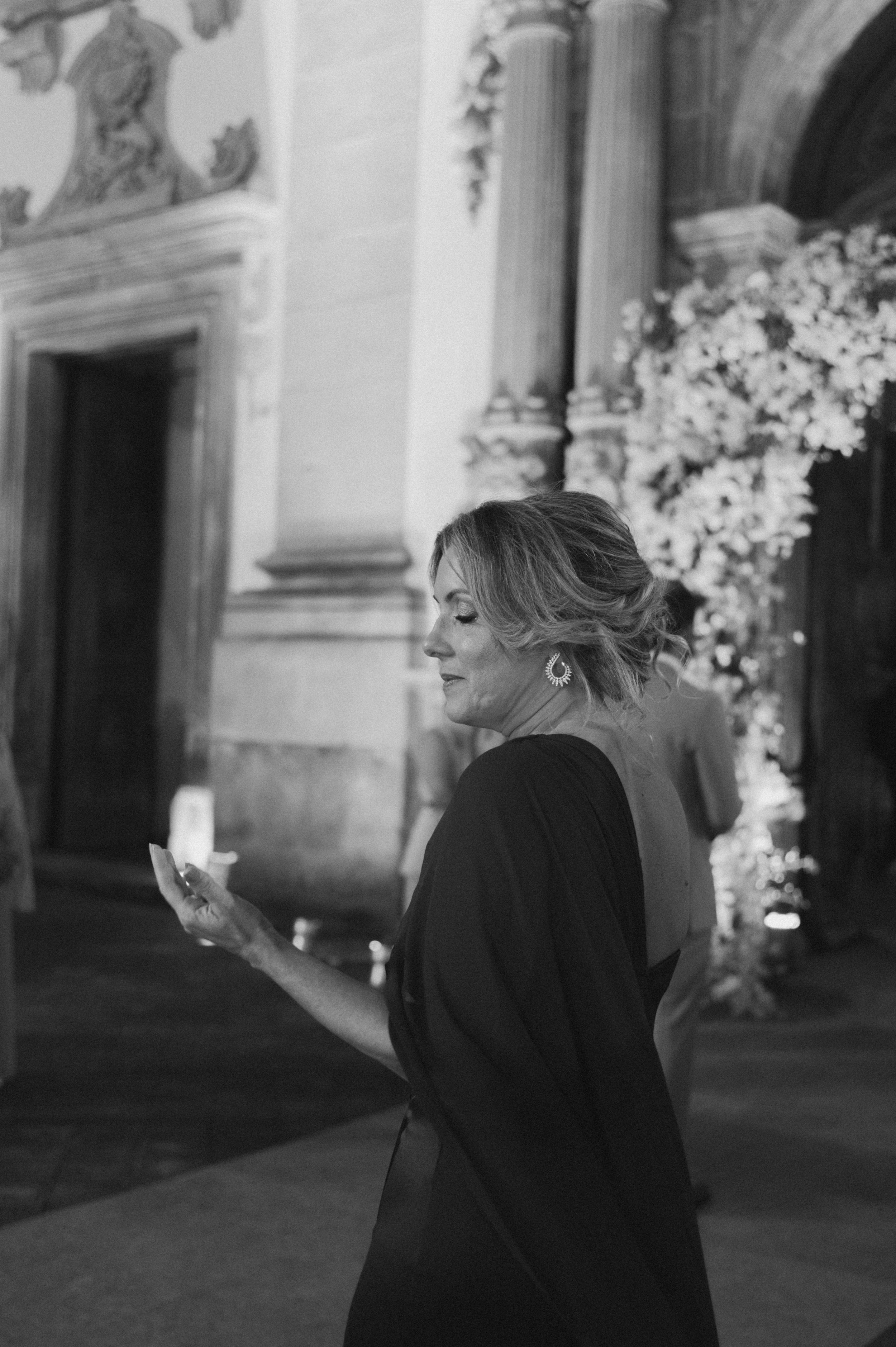 A person in a black dress holds a candle while standing near ornate architectural columns in a black and white photo.