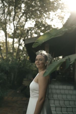 A bride in a white dress and vintage-style veil stands against a brick wall as sunlight filters through trees.
