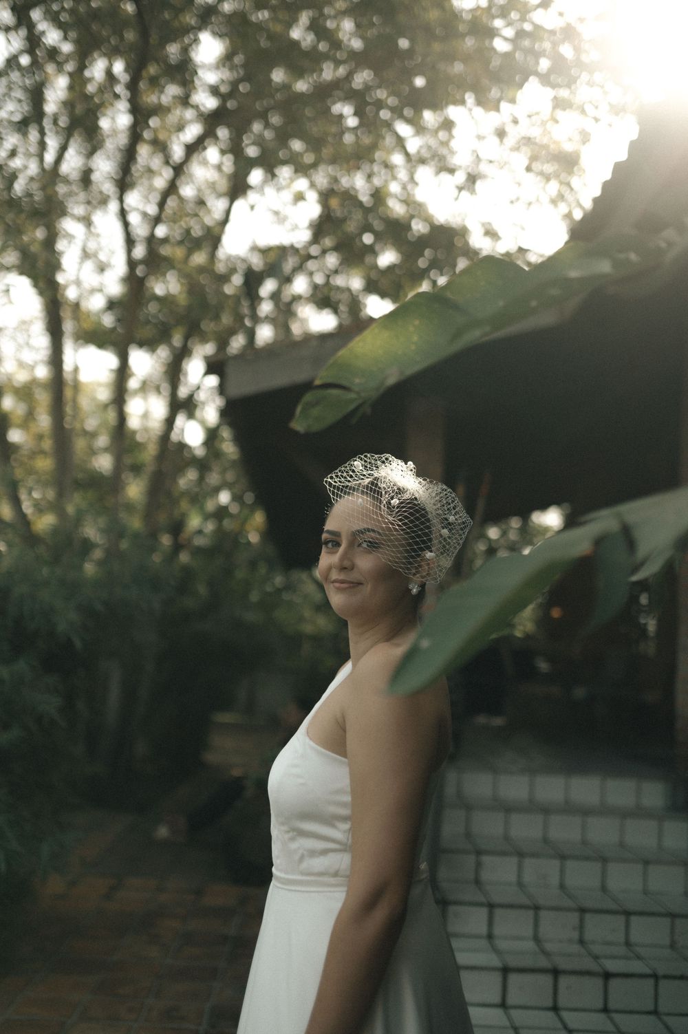 A bride in a white dress and vintage-style veil stands against a brick wall as sunlight filters through trees.