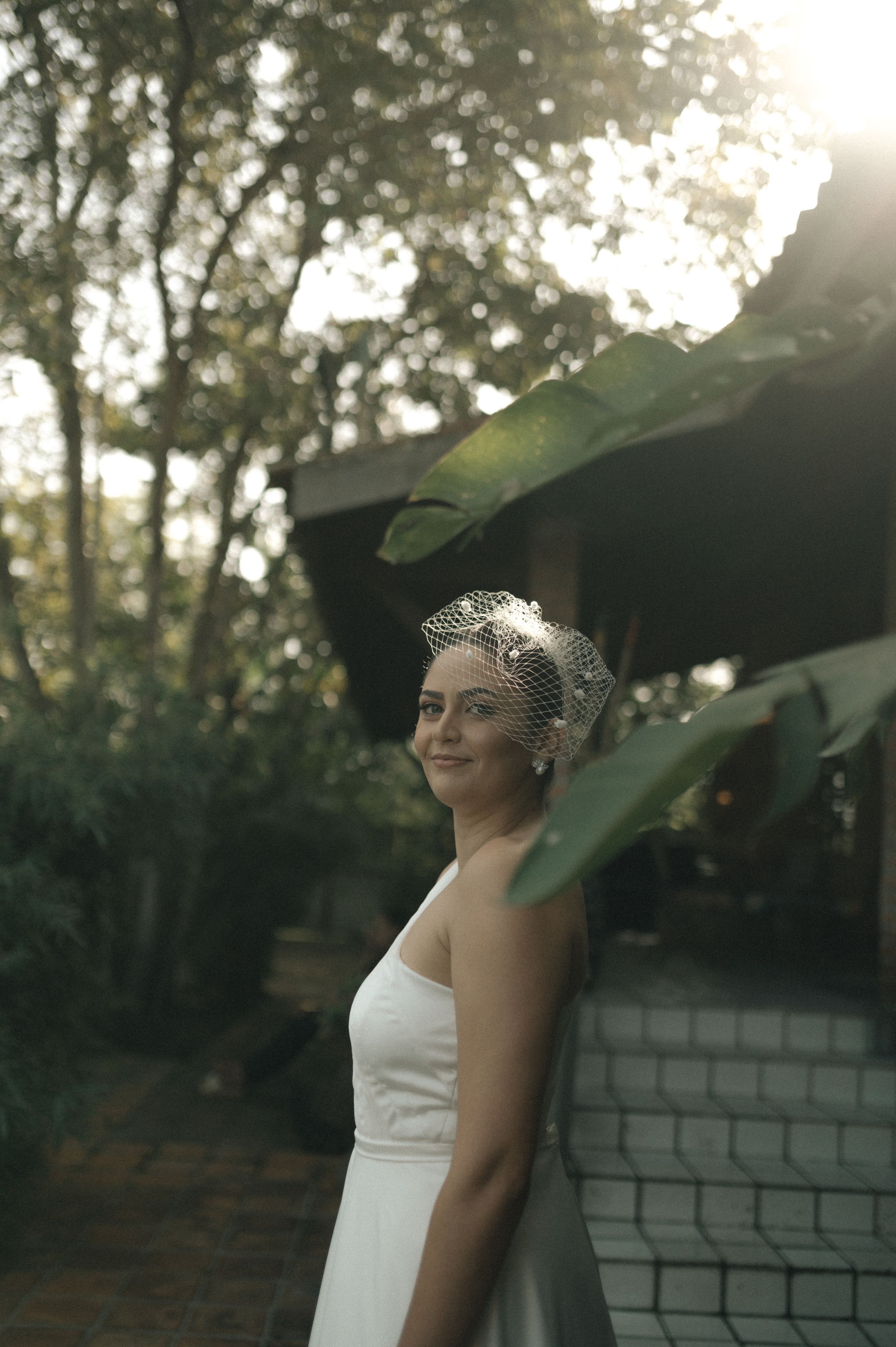 A bride in a white dress and vintage-style veil stands against a brick wall as sunlight filters through trees.
