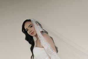 Bride wearing a white gown and lace-trimmed veil, posing elegantly against a soft neutral background.