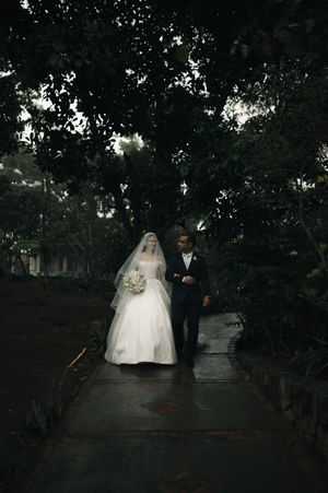A romantic wedding couple walks together under a canopy of dark trees in an evening garden setting.