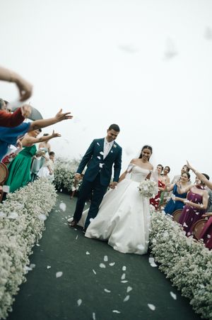 Wedding ceremony guests toss flower petals as couple walks down outdoor aisle with white floral decorations.