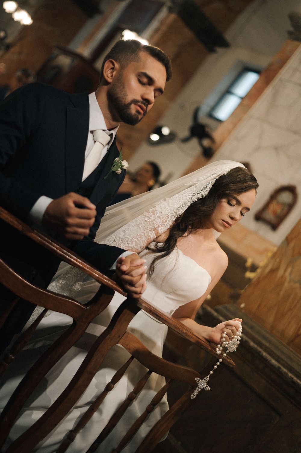 Bride and groom praying together at church altar during wedding ceremony, bride holding rosary beads.