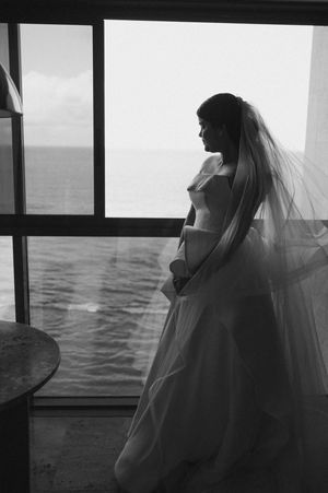 Black and white photo series of a bride in a flowing veil gazing out over the ocean through large windows.