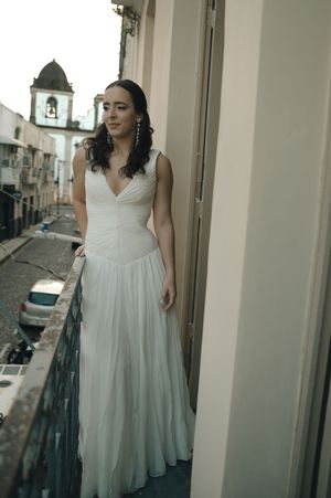 A bride in a flowing white wedding dress poses elegantly on a balcony overlooking a European street.