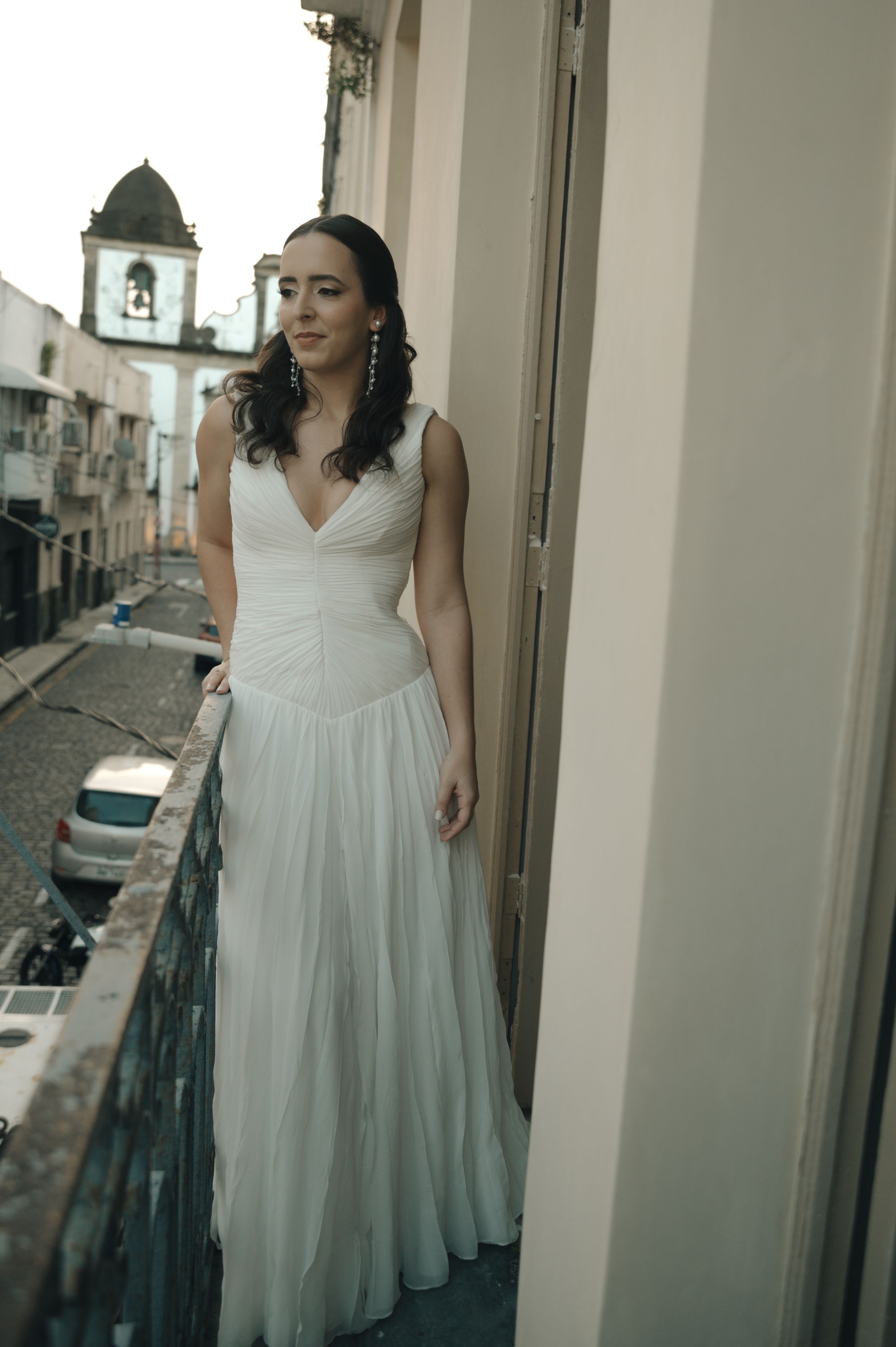 A bride in a flowing white wedding dress poses elegantly on a balcony overlooking a European street.