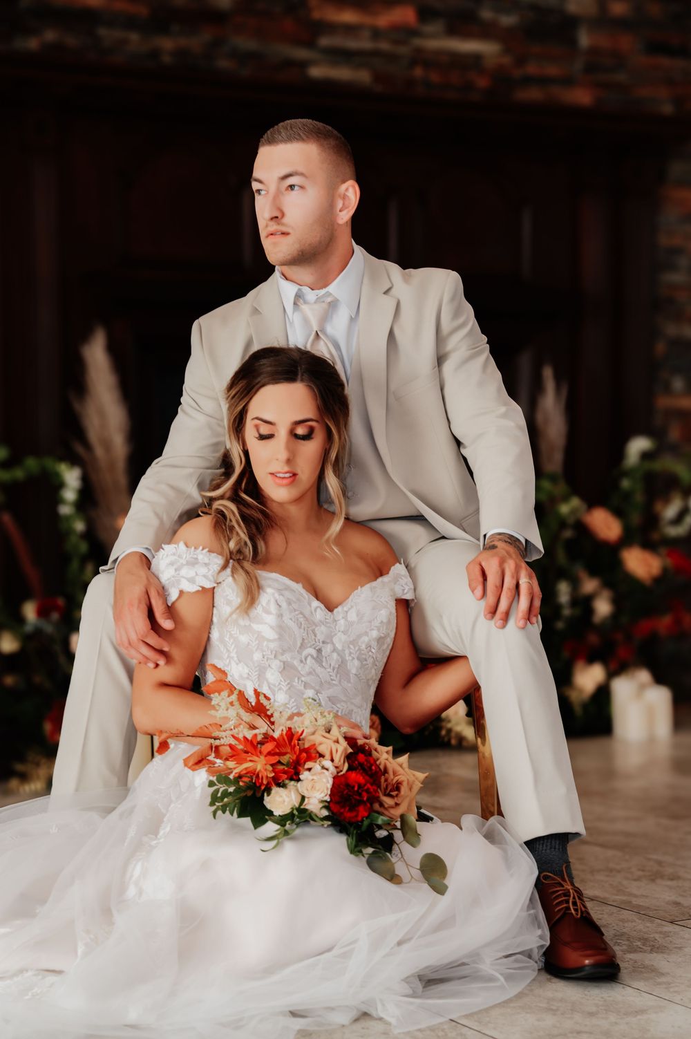 Bride and groom posing elegantly indoors, she in a lace off-shoulder gown with a vibrant bouquet, he in a tan suit.