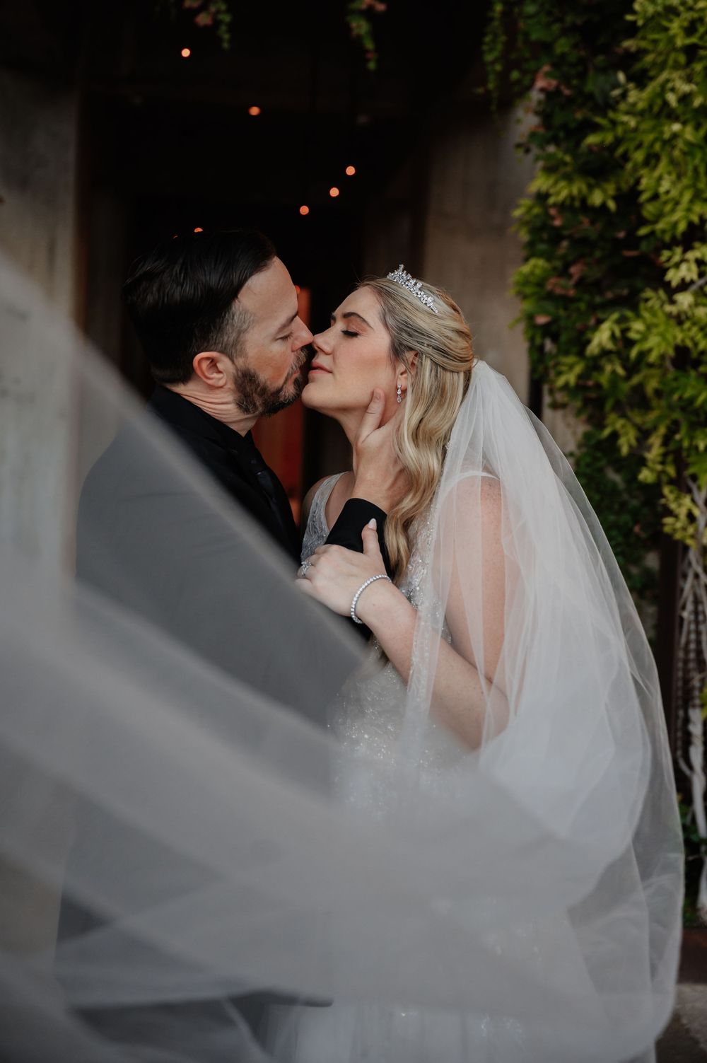 Bride and groom sharing a passionate kiss outdoors with a flowing veil and string lights backdrop.