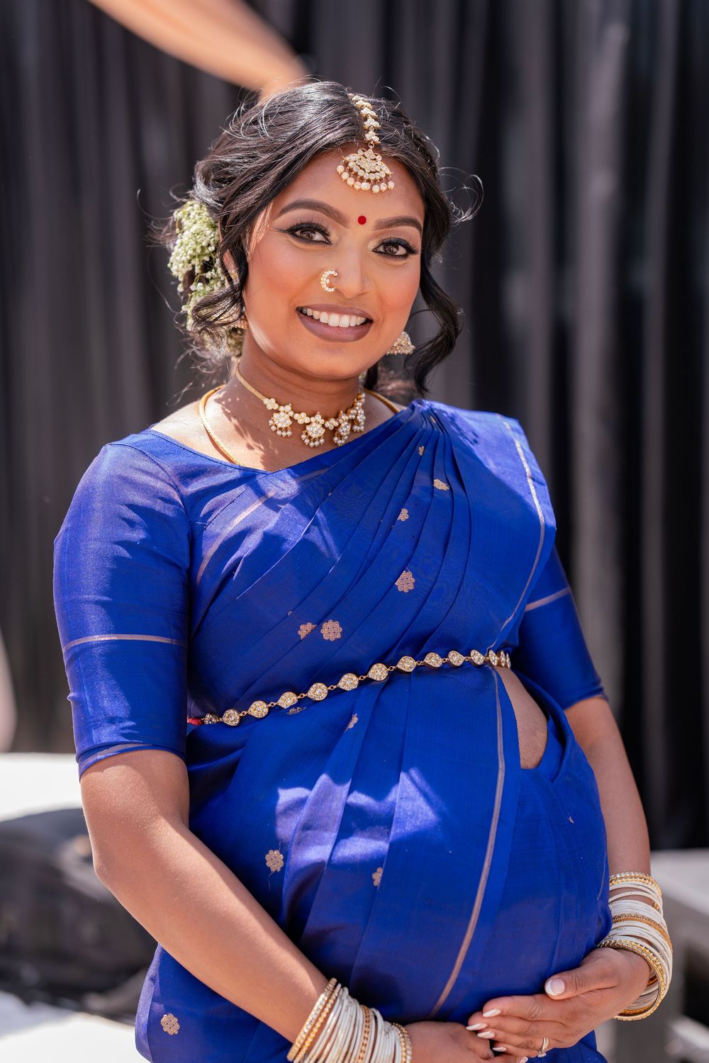 Person wearing royal blue traditional Indian attire with gold accessories against dark backdrop.
