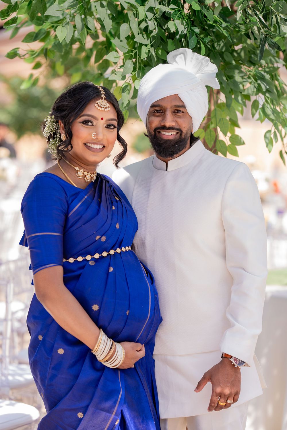 Couple expecting their first baby in traditional Indian wedding attire posing under green foliage.