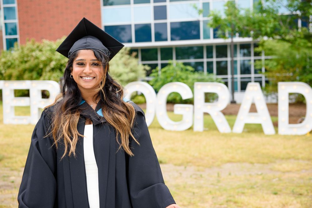 Graduate in cap and gown smiling in front of grad sign on campus.