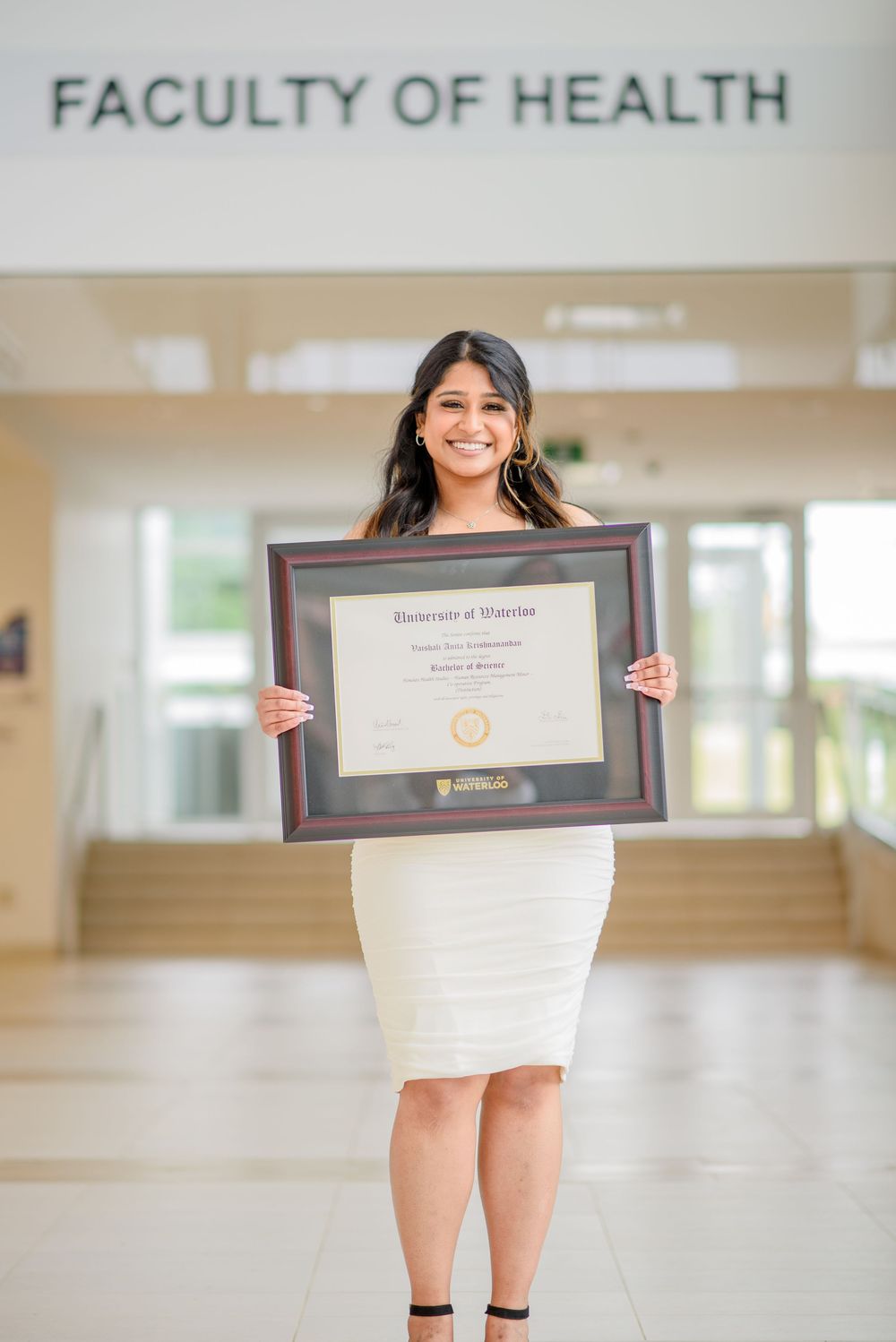 Graduate in white dress holding framed diploma at the Faculty of Health.