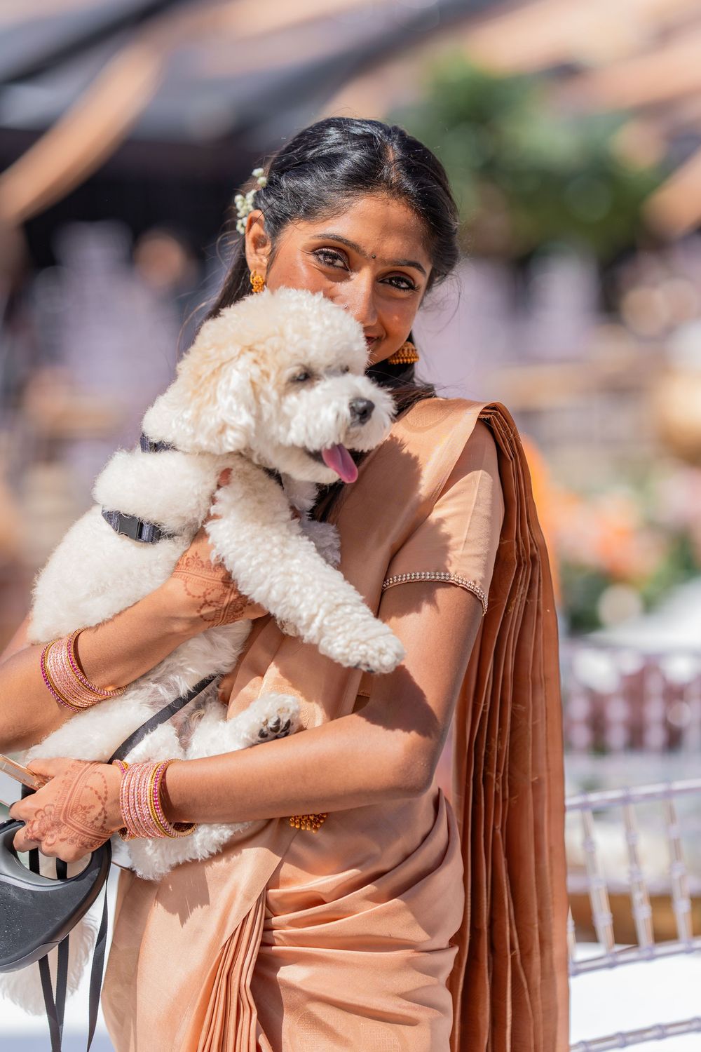 Woman wearing peach colored saree embraces small white dog outdoors.
