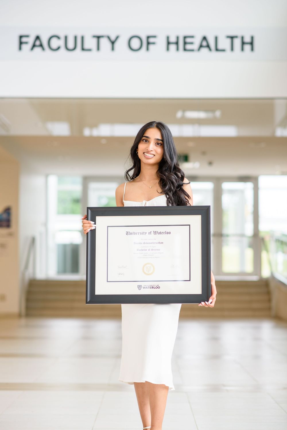 Graduate showing her framed diploma in front of Faculty of Health sign.