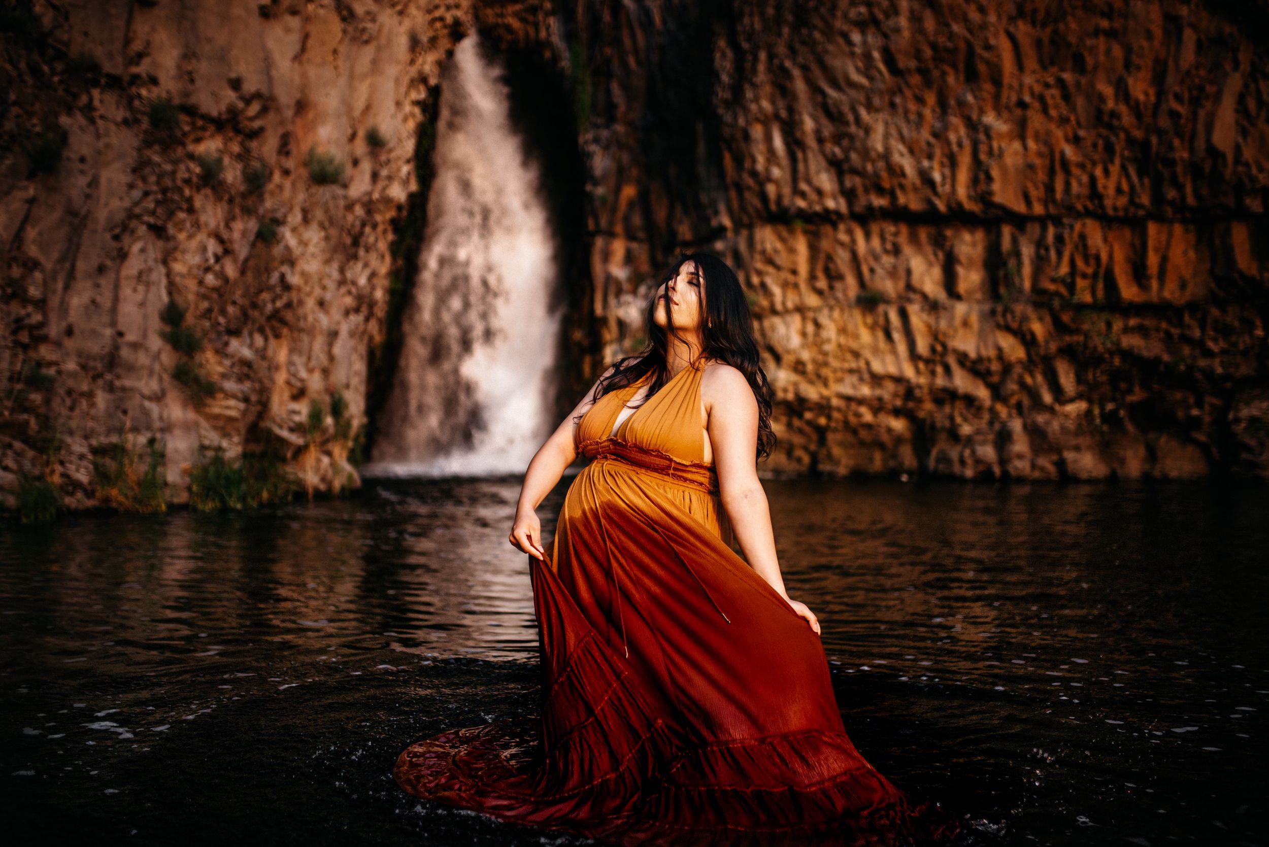 Maternity Mother with an orange twirling dress as she stands in front of a waterfall in Spokane, Washington.