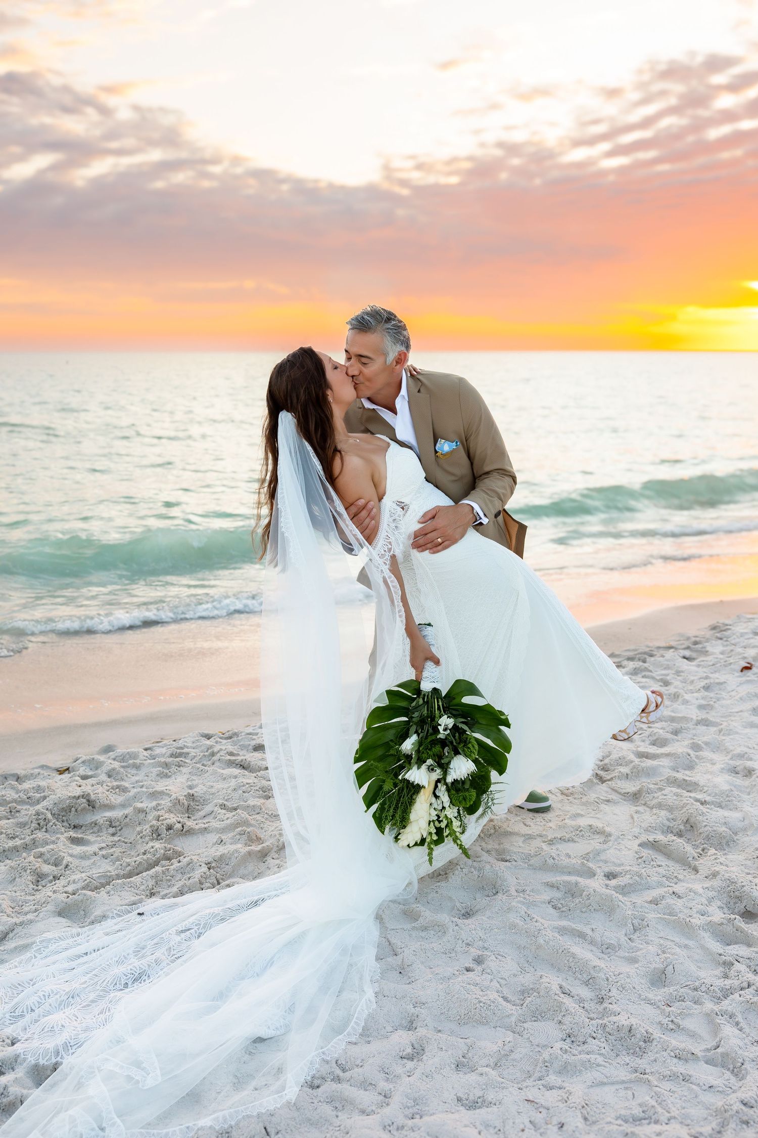 Bride in lace gown and groom in purple tuxedo pose before ocean mural at wedding.