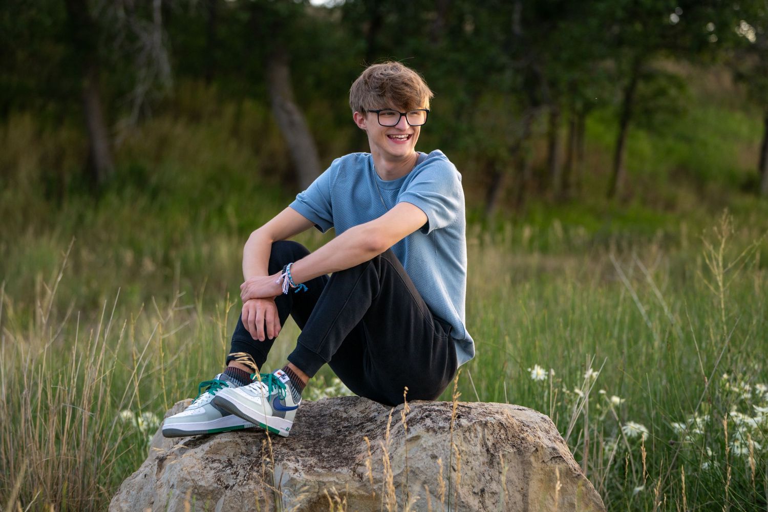 High school senior sits casually on rock in grassy field wearing blue t-shirt and sneakers.