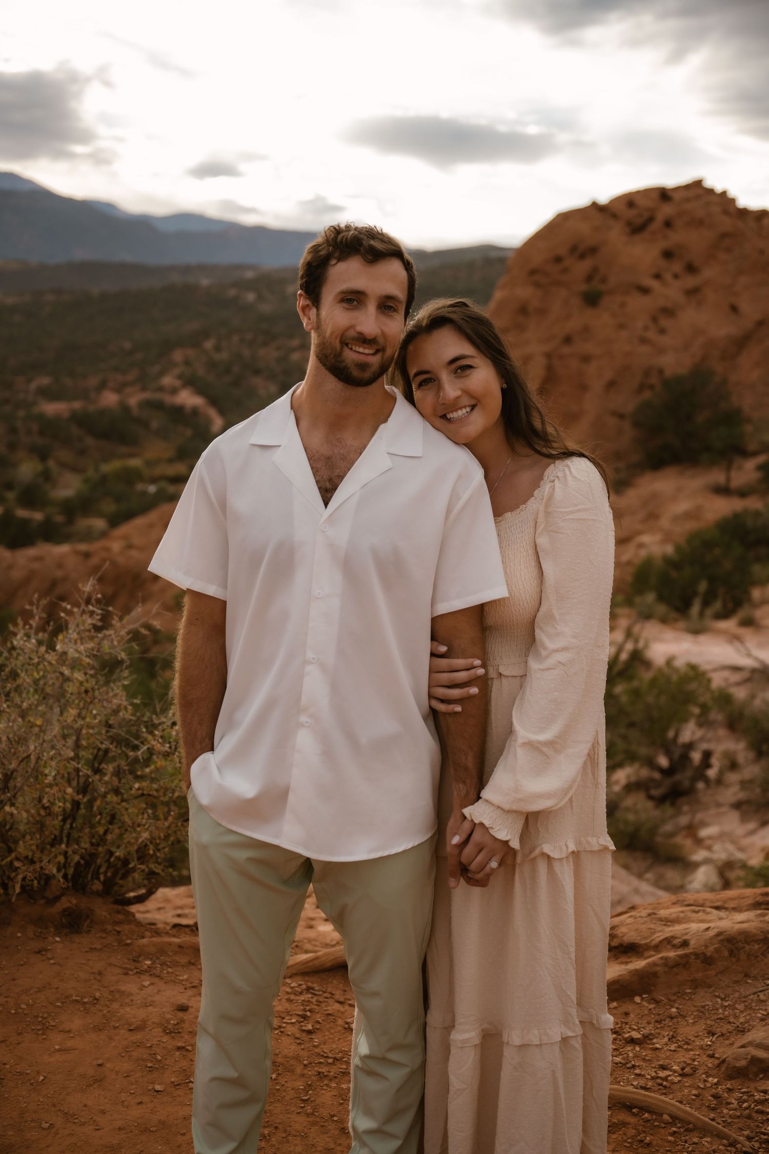 Couple in white outfits embracing against a scenic desert mountain backdrop during sunset.