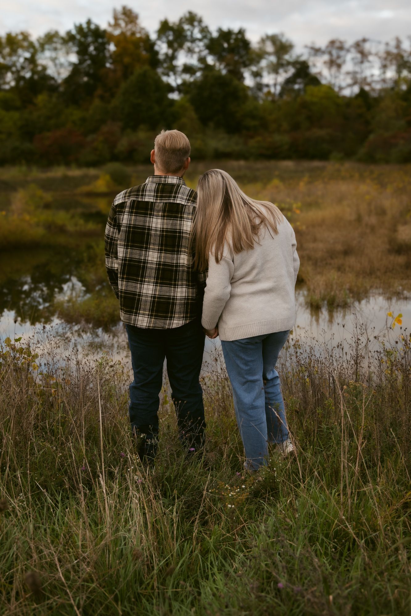 A couple in casual attire stands together in a scenic field of tall grass and wildflowers at sunset.
