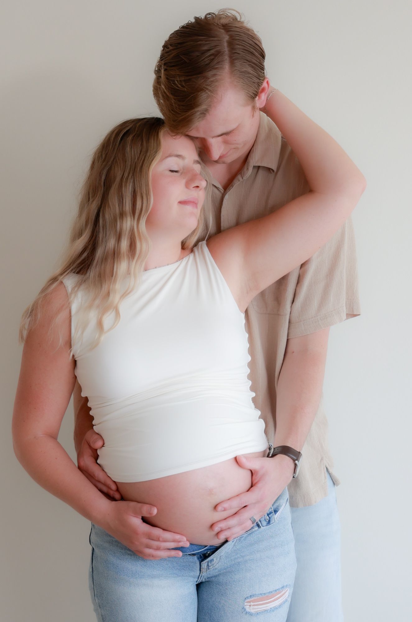 Intimate studio maternity photos showing a couple embracing in a white crop top and beige shirt.