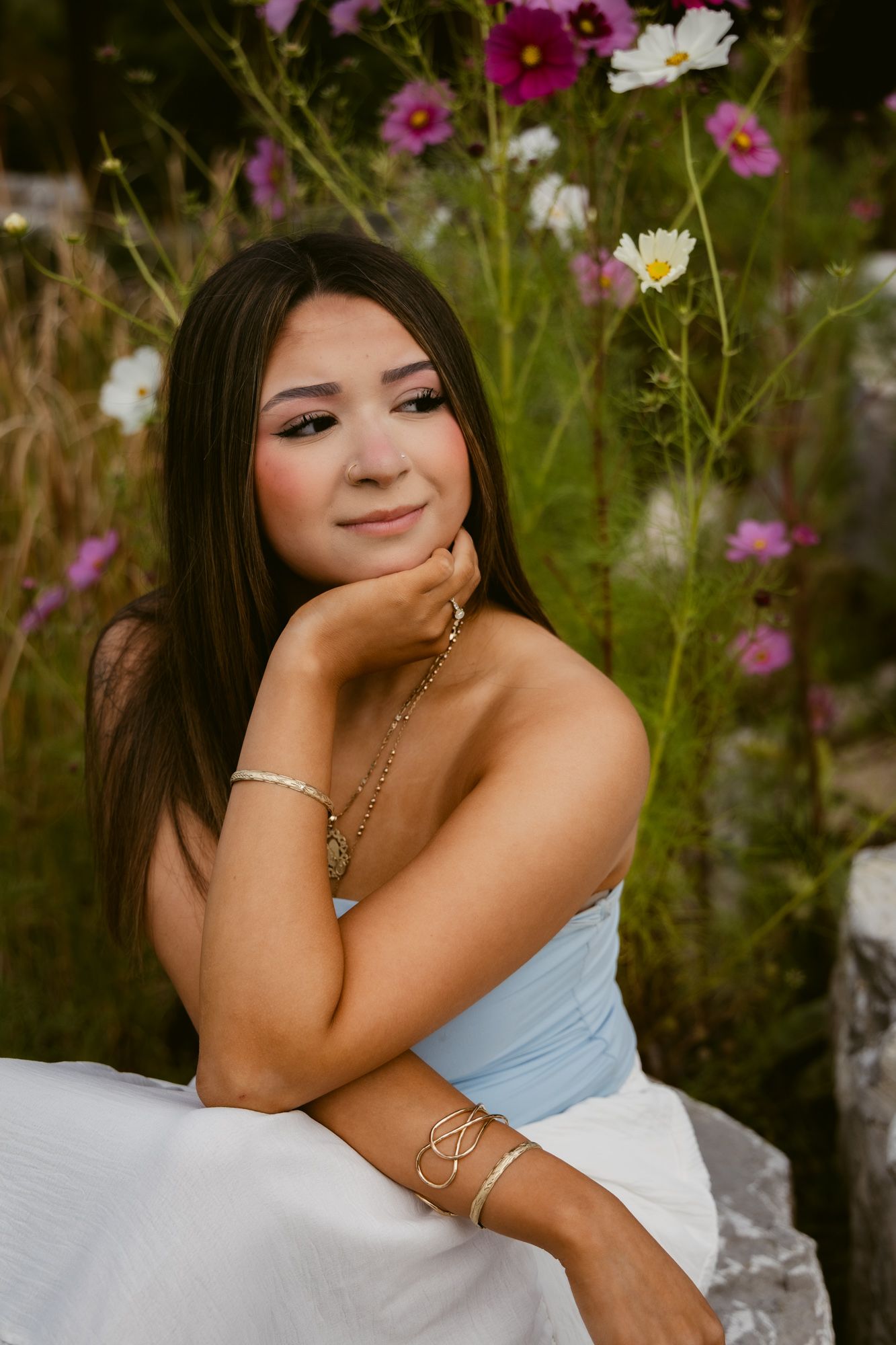 A person in a light blue dress poses outdoors among pink and white cosmos flowers in a garden setting.