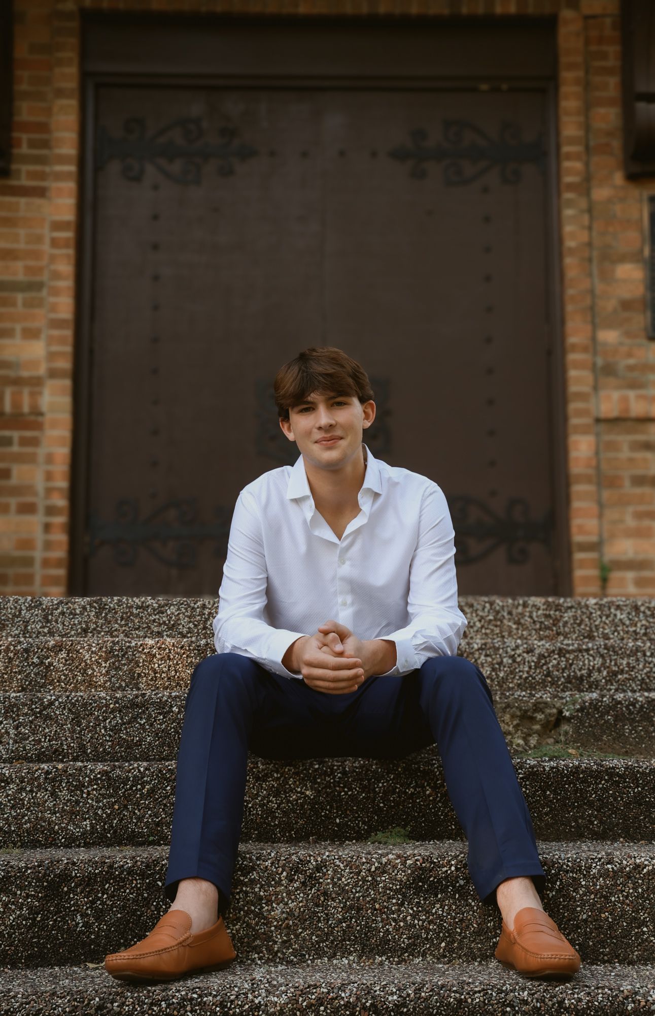 Person in white shirt and blue pants sits casually on concrete steps against brick wall backdrop.