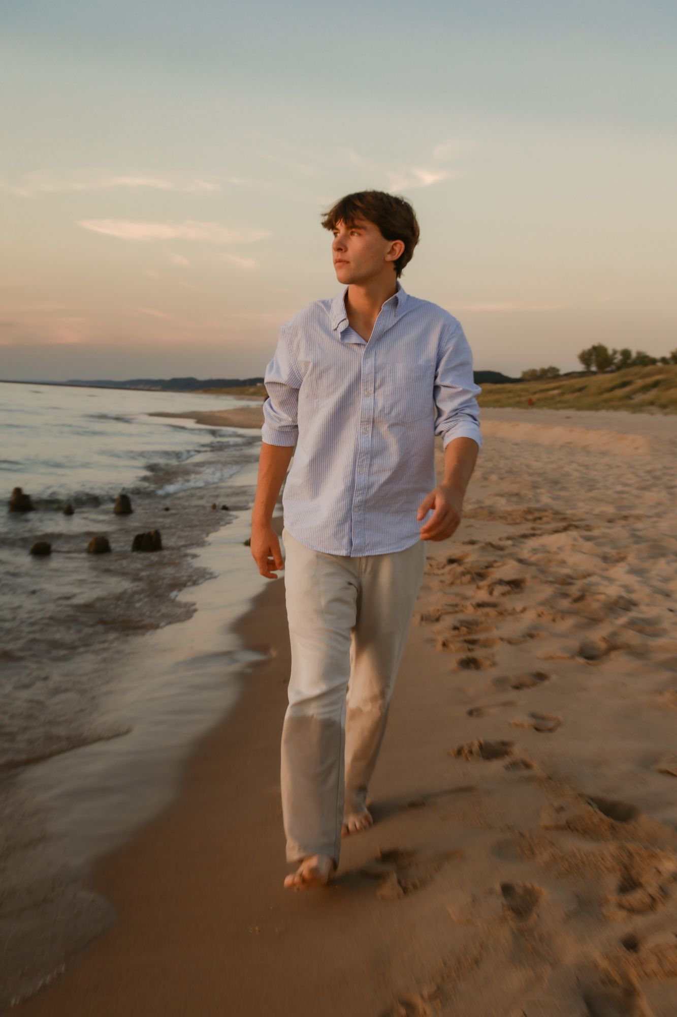 A person in a light blue button-down shirt and khakis walks barefoot along a beach at sunset.