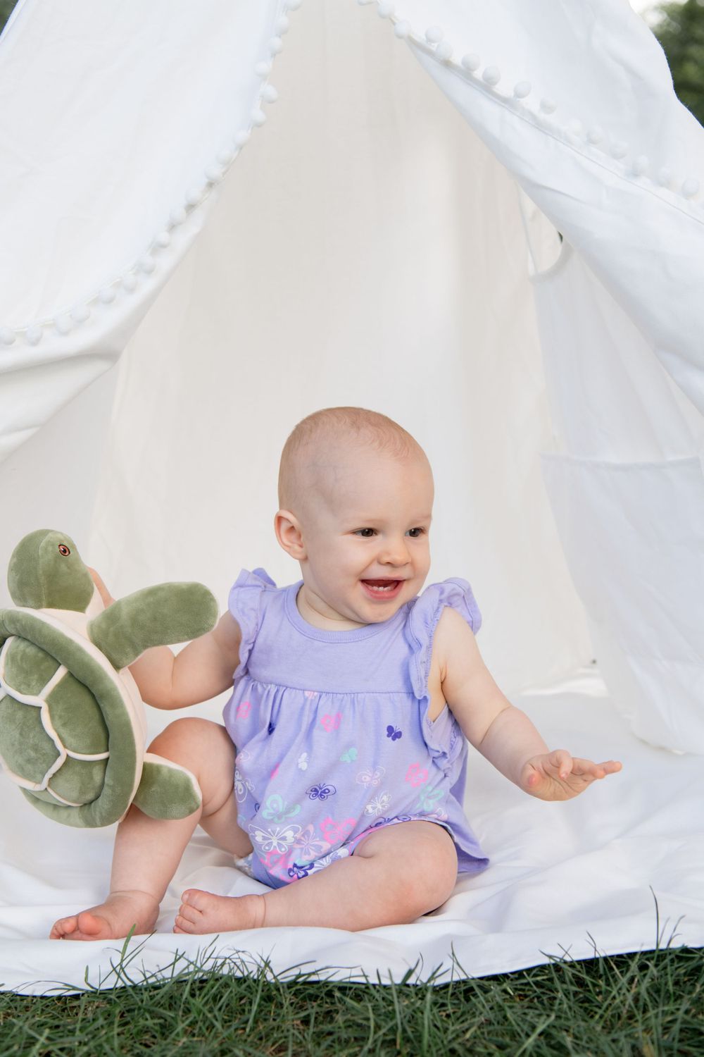 Purple-dressed baby playing with plush turtle in white teepee tent outdoors.