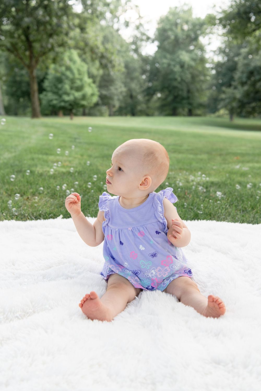Baby in purple floral romper sits on white blanket outdoors while reaching for bubbles in a sunny garden setting.
