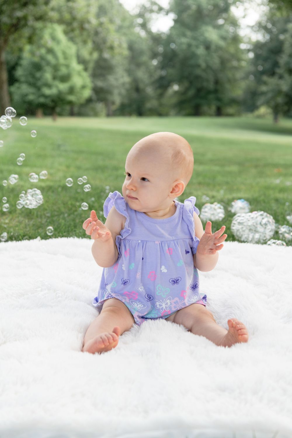 Baby wearing lavender outfit sits on white blanket playing with soap bubbles in a green park setting.