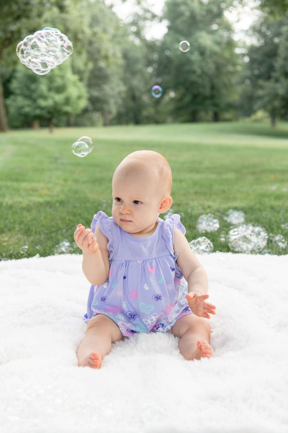 Baby in ruffled purple outfit sits on white blanket reaching for floating bubbles against green background.