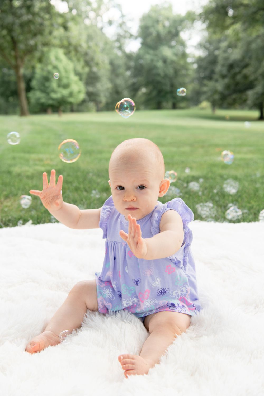 Small baby in purple dress plays with bubbles while sitting on white blanket outdoors.