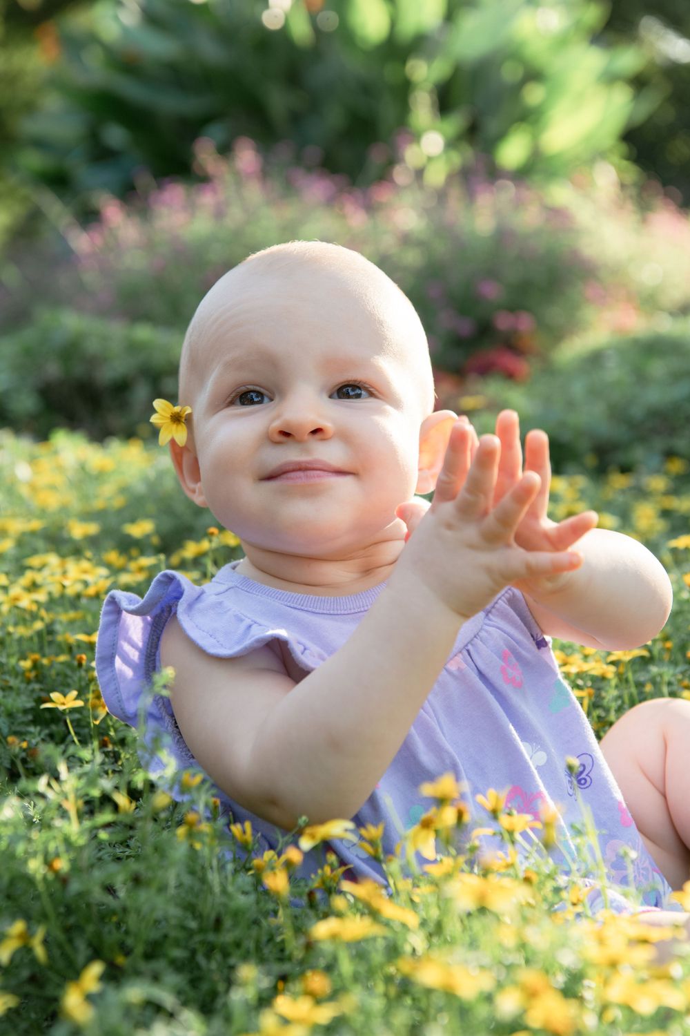 Baby in lavender outfit plays among yellow flowers in blooming garden.