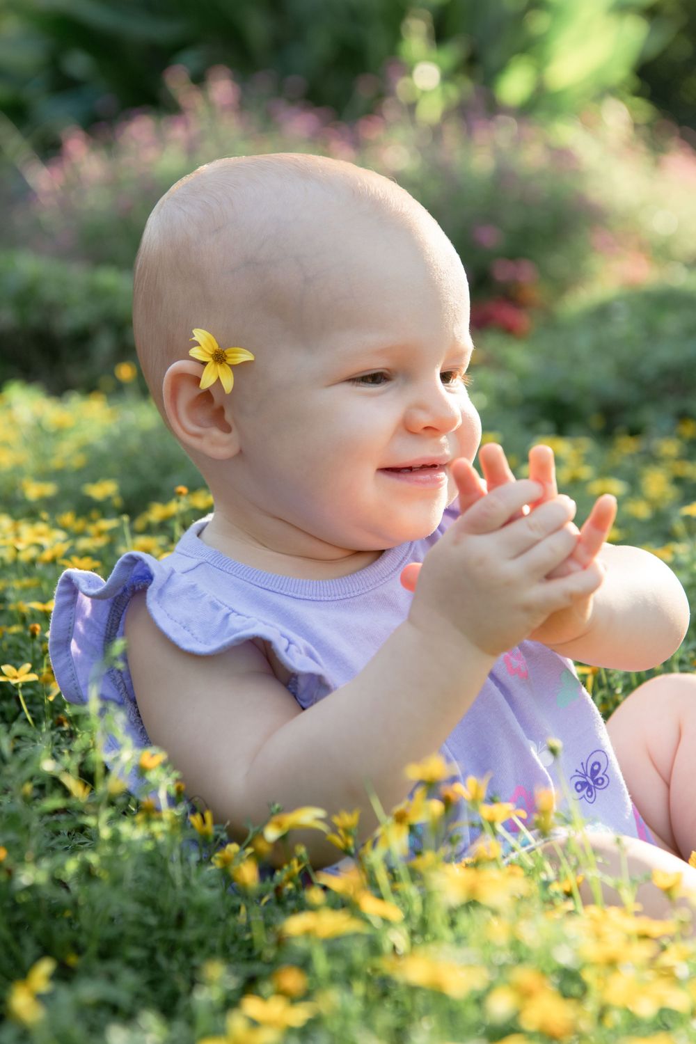 Baby with yellow flower plays in garden of blooming flowers while wearing purple dress.