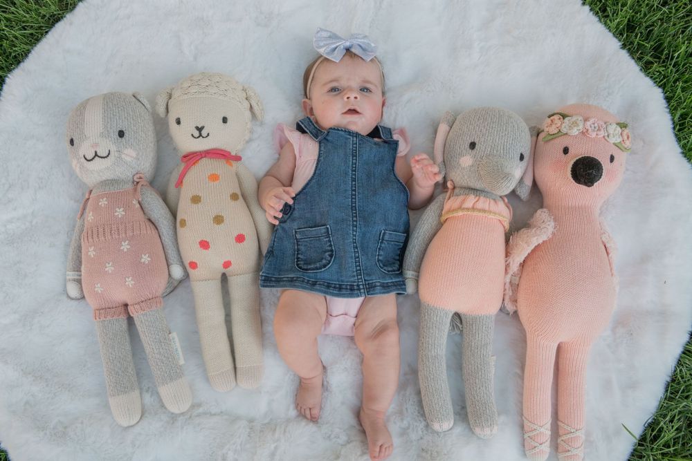 Baby in denim outfit lying on white blanket surrounded by plush pink and beige stuffed animals.